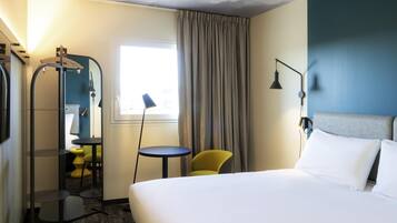 A hotel room with a bed, a chair, a small table, a mirror, and a window with curtains.