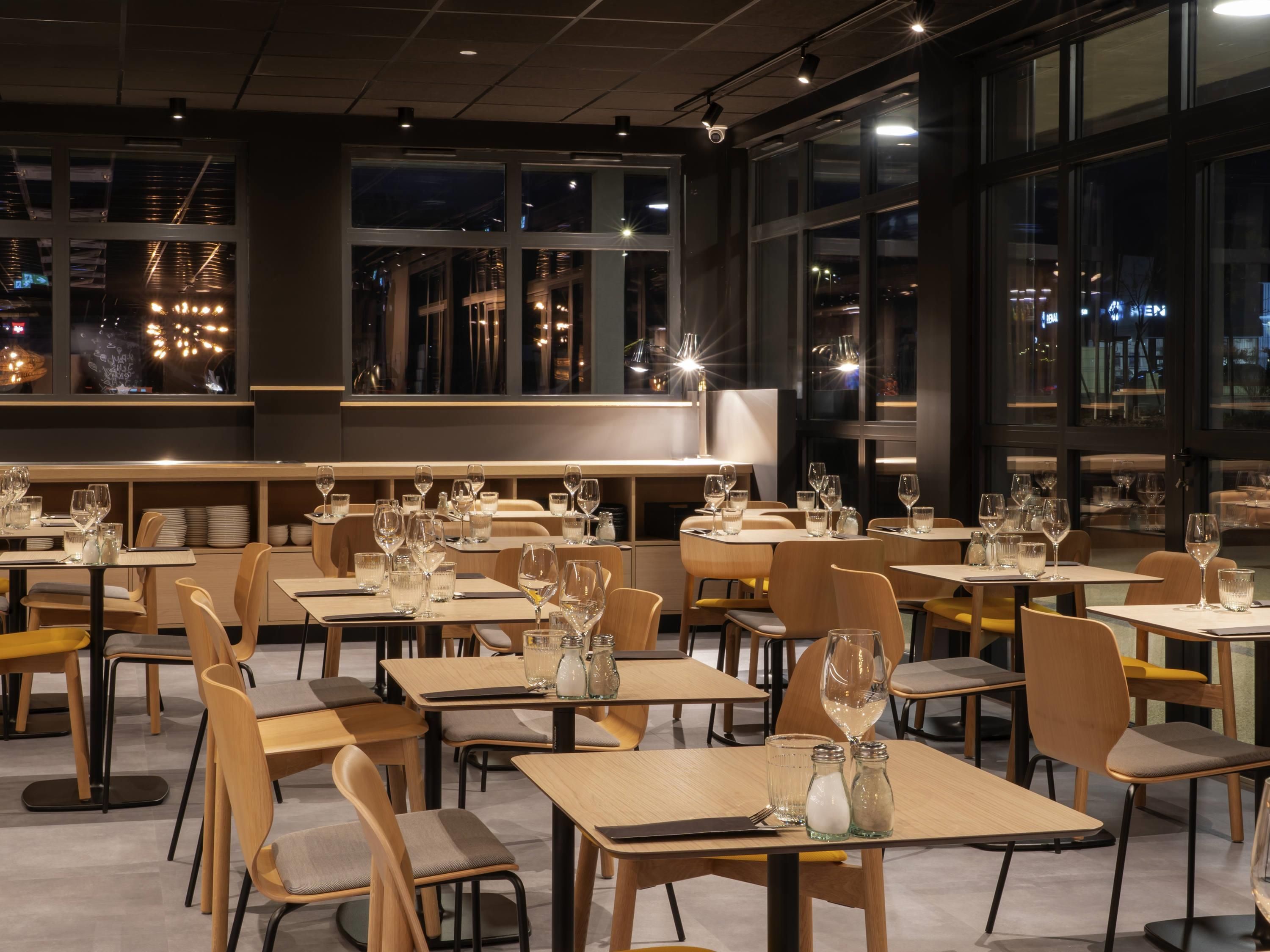A restaurant interior with tables set for dining, large windows, and a city view at night.