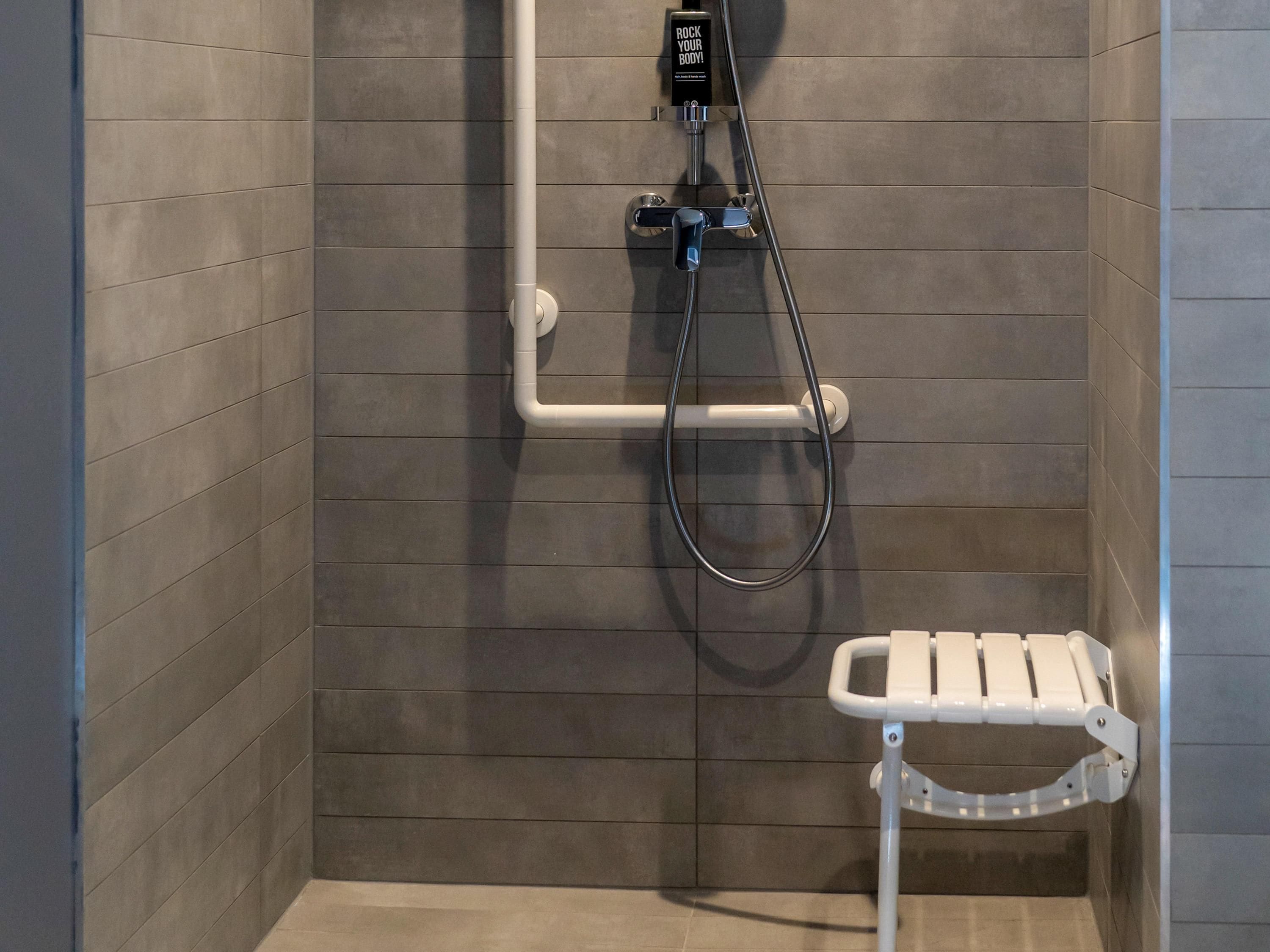 A shower area with a handheld showerhead, a wall-mounted control panel, and a white grab bar.