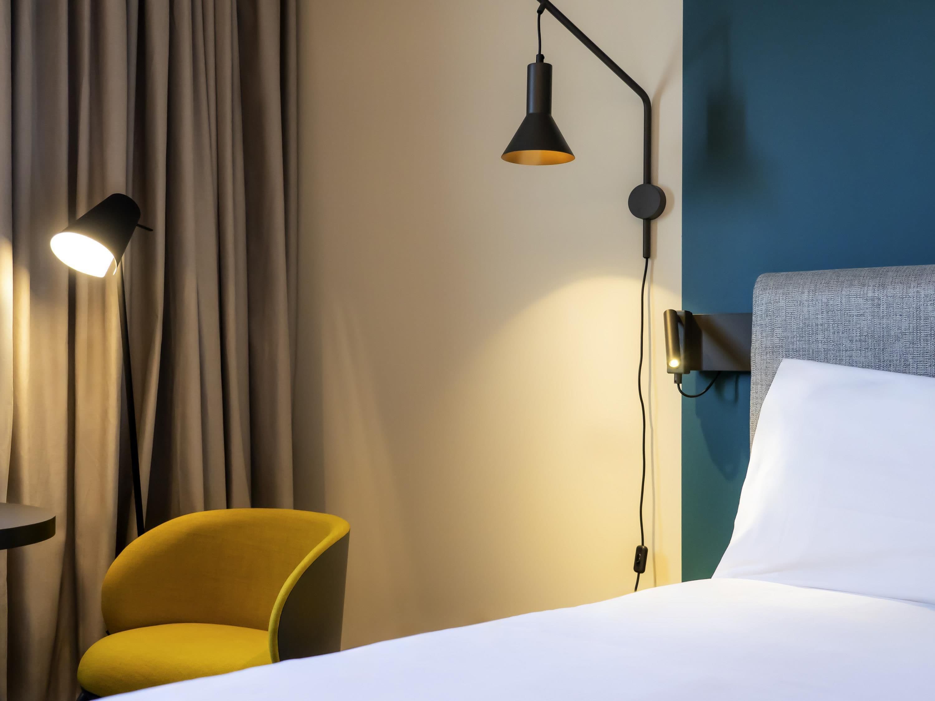 Quarto de hotel com cama, uma poltrona amarela, luminária na parede e cortina.