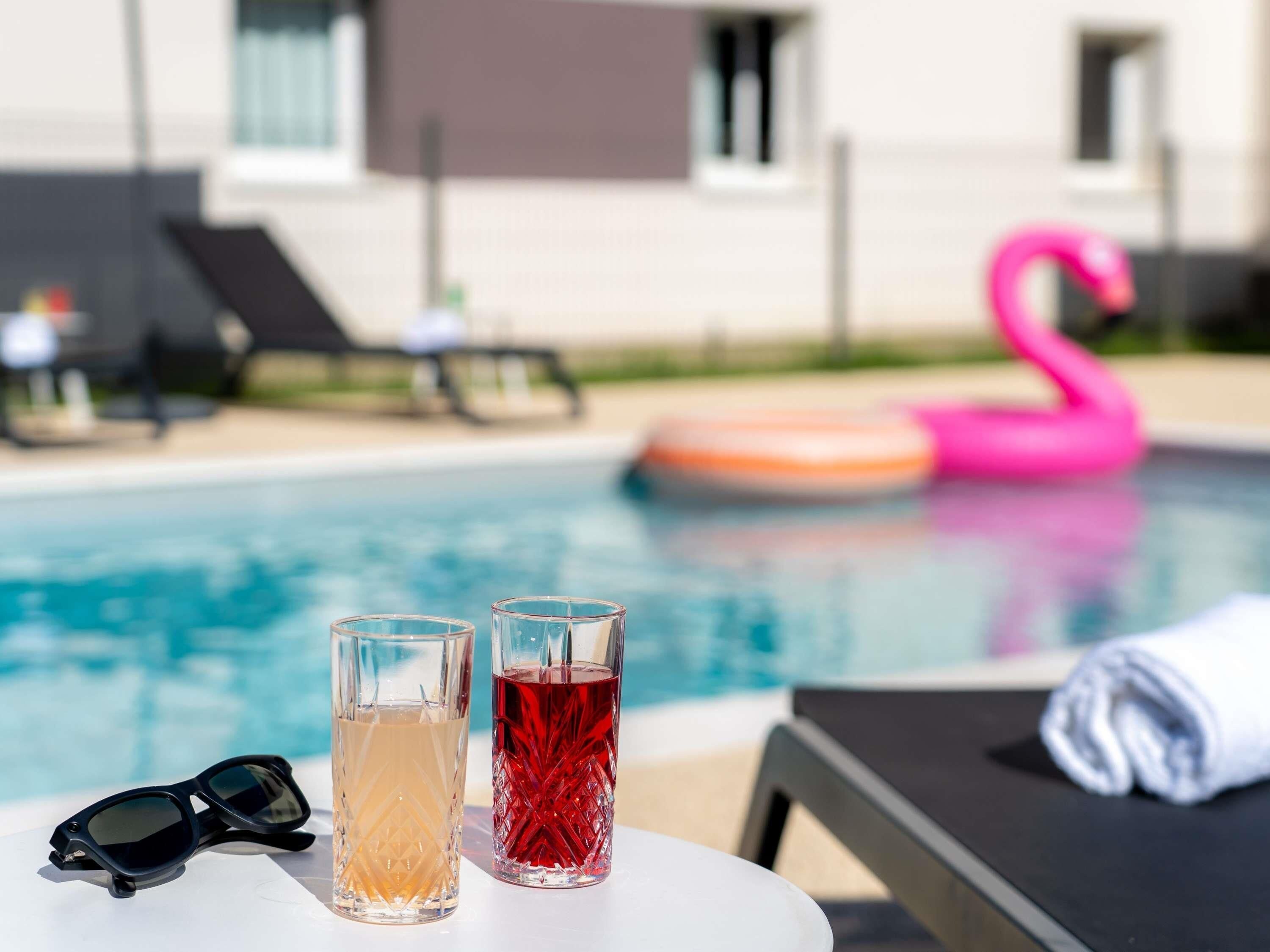 Una mesa junto a la piscina con dos vasos de bebidas, un par de gafas de sol y un flotador con forma de flamenco rosa.