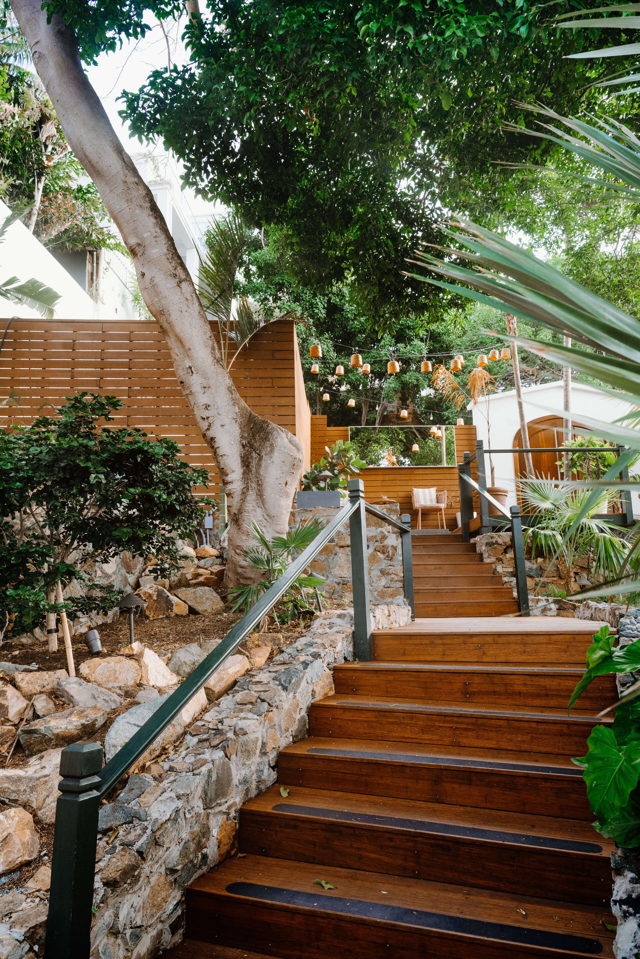 Un escalier en bois, avec des murs en pierre et une rampe en métal, entouré de verdure et délimité par une clôture en bois.
