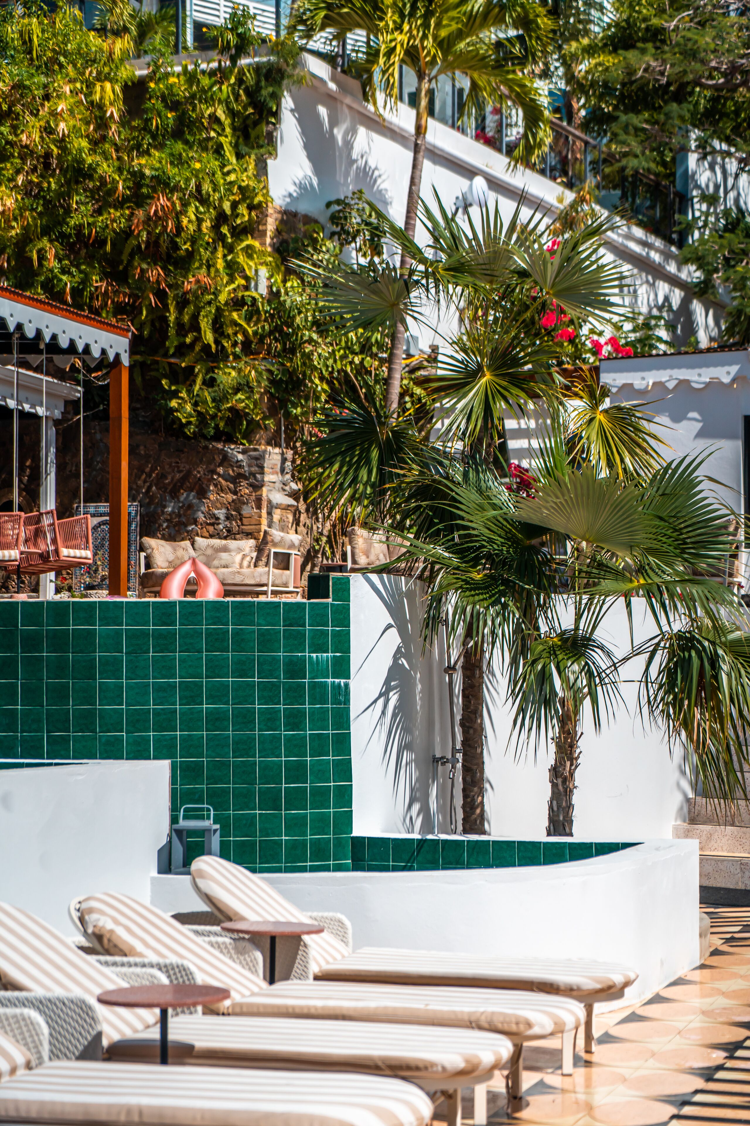 Un espace aménagé au bord de la piscine, avec des chaises longues, un mur recouvert de carreaux et des palmiers.