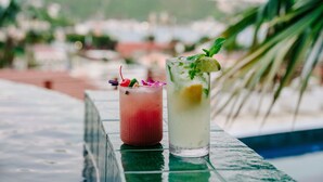 Two drinks on a table with a pool and palm trees in the background.