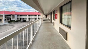 A hotel corridor with a balcony, a red-roofed building in the background, and a parking lot with cars.