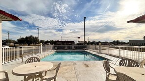 A rooftop pool area with a view of the city skyline, surrounded by white railings and furnished with plastic chairs and tables.