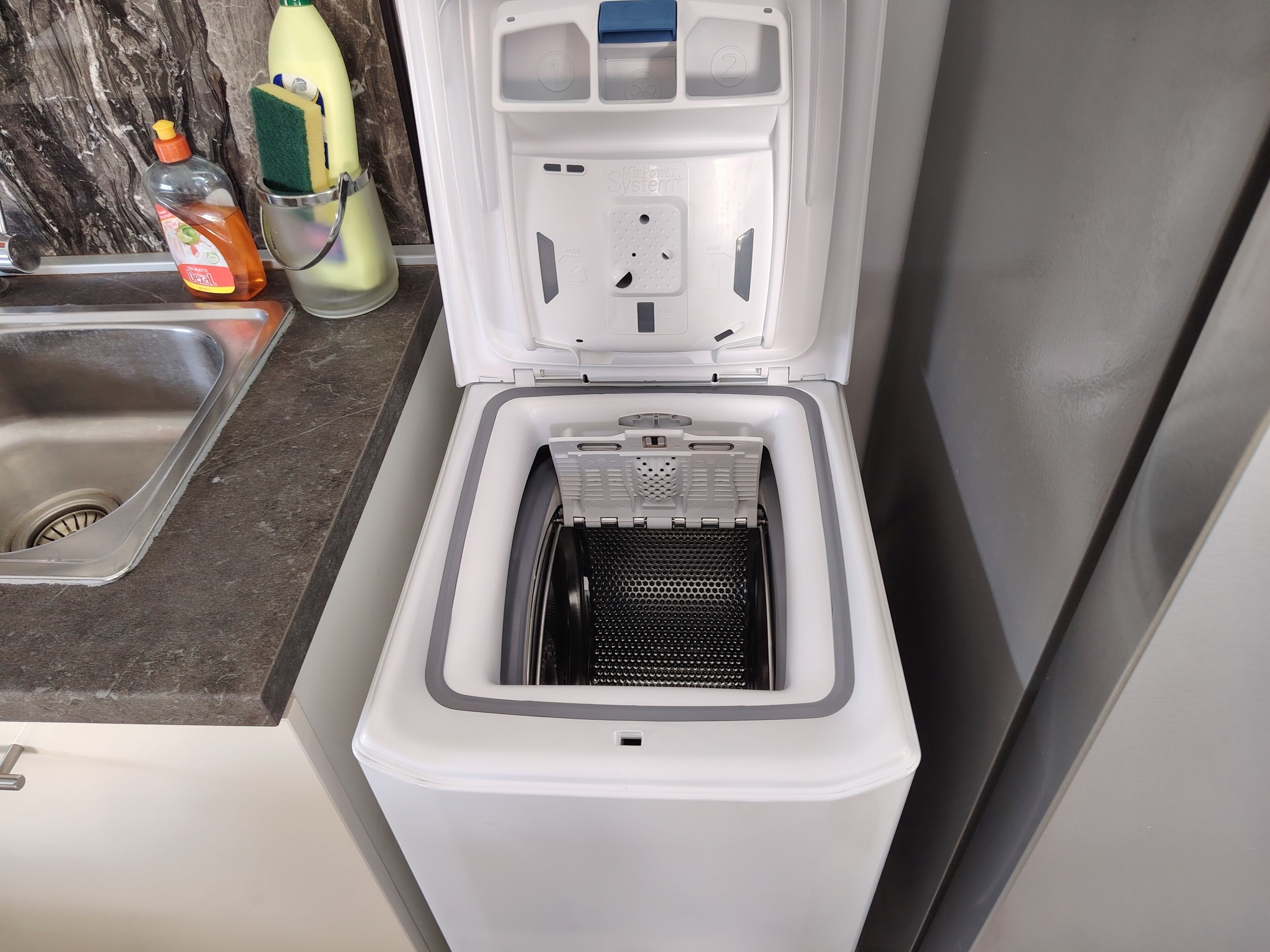 Een compact, wit apparaat met ingebouwde ijsmachine, geplaatst in een keuken met een marmeren achterwand en een roestvrijstalen spoelbak.