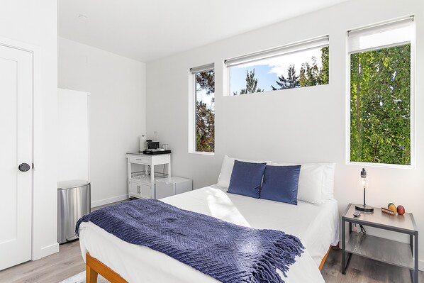 A modern bedroom with a large bed, a bedside table, a small desk, and a trash bin.