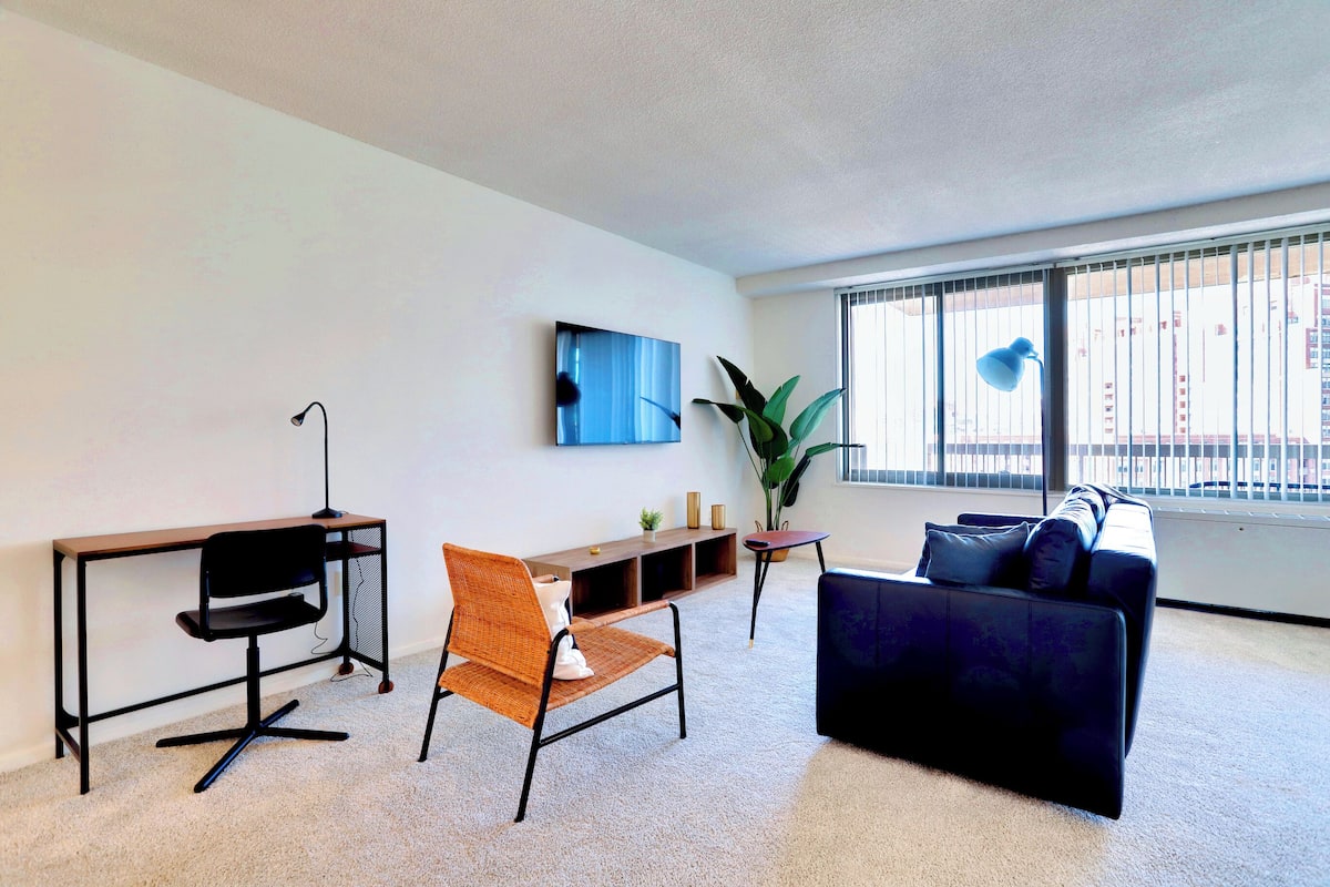 Un salon moderne comprenant un bureau, deux fauteuils, un canapé, une grande fenêtre donnant sur des immeubles et une plante en pot.