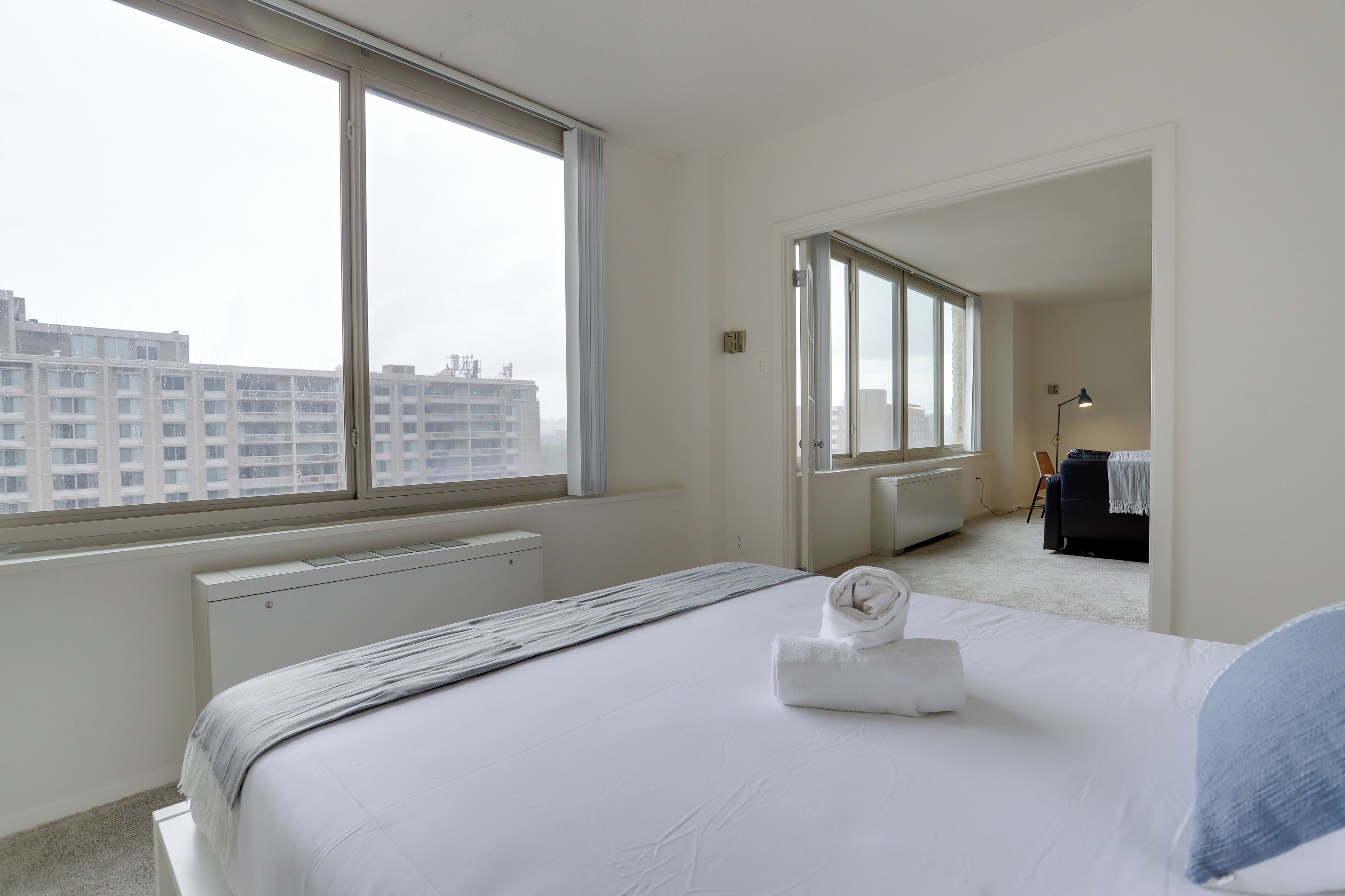 A hotel room with a large window, a bed with a white duvet, and a radiator.