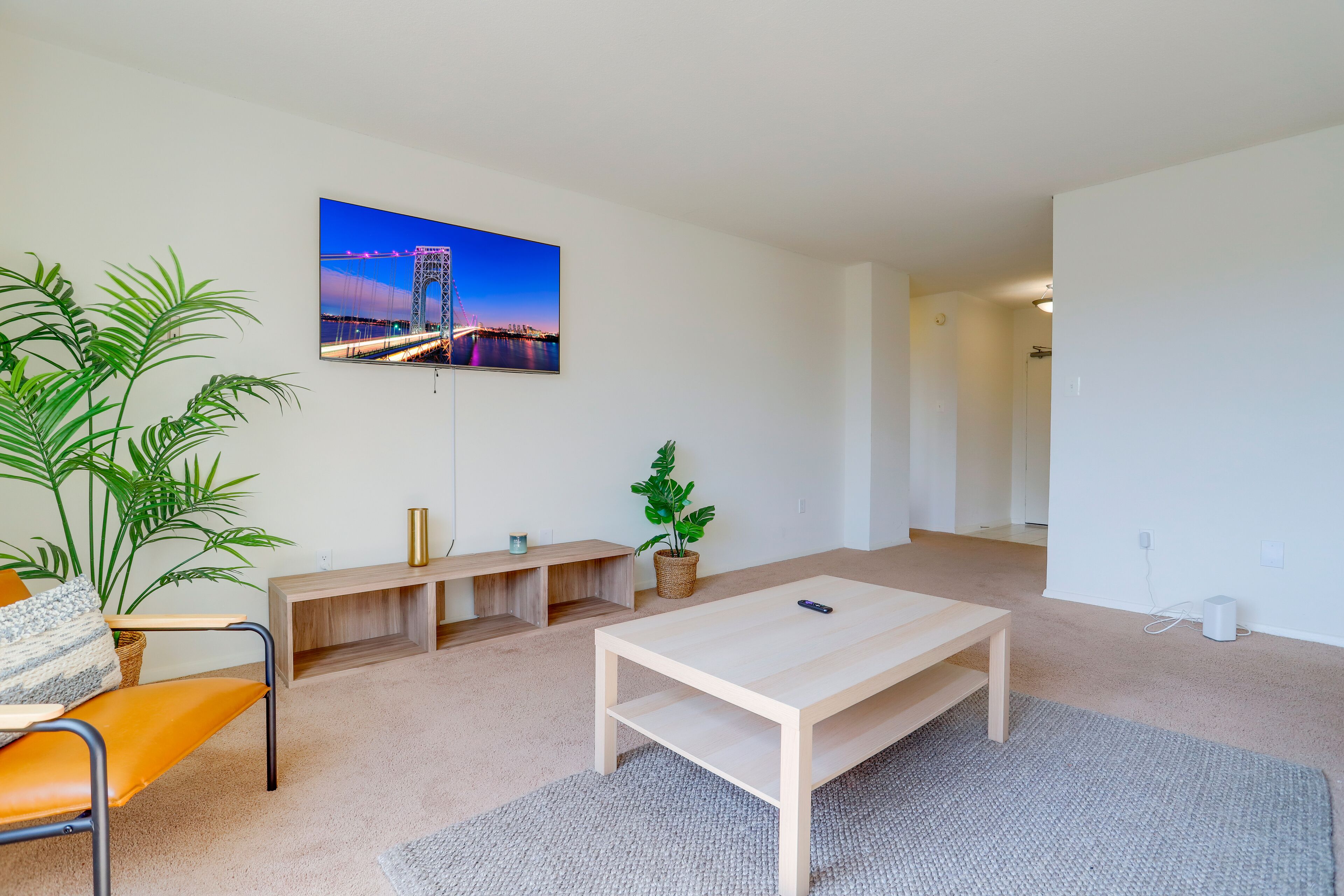 Uma sala de estar moderna com uma grande televisão de tela plana na parede, uma mesa de centro de madeira, um sofá, um banquinho e duas plantas em vasos.