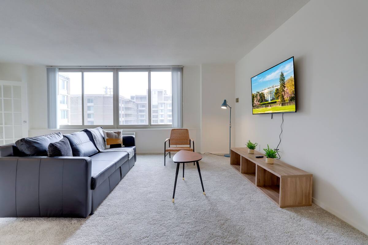 Un salon moderne avec un canapé gris, une table basse en bois, un fauteuil, un téléviseur fixé au mur et une grande fenêtre donnant sur la ville.