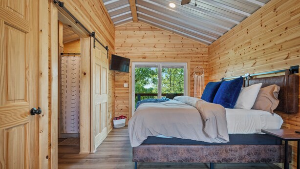 A wooden cabin bedroom with a bed, a television, and a window with a view of trees.