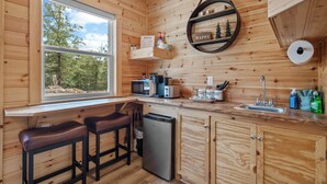 A wooden cabin interior with a kitchen area, including a sink, microwave, and refrigerator. There are two stools and a window with a view of trees.