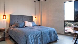Ett modernt sovrum med en stor säng, två sänglampor, utsikt över naturen genom ett fönster och ett litet bord med en stol.