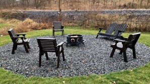 Un espace extérieur aménagé avec cinq fauteuils Adirondack et un foyer, entouré de gravier et d’herbe, avec une colline boisée en arrière-plan.