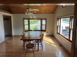 Un comedor amplio con una gran mesa de madera y sillas, un ventilador de techo y ventanas grandes con vistas al exterior.