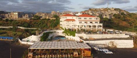 A beachfront hotel with multiple balconies, a covered outdoor seating area, and a view of the surrounding town.