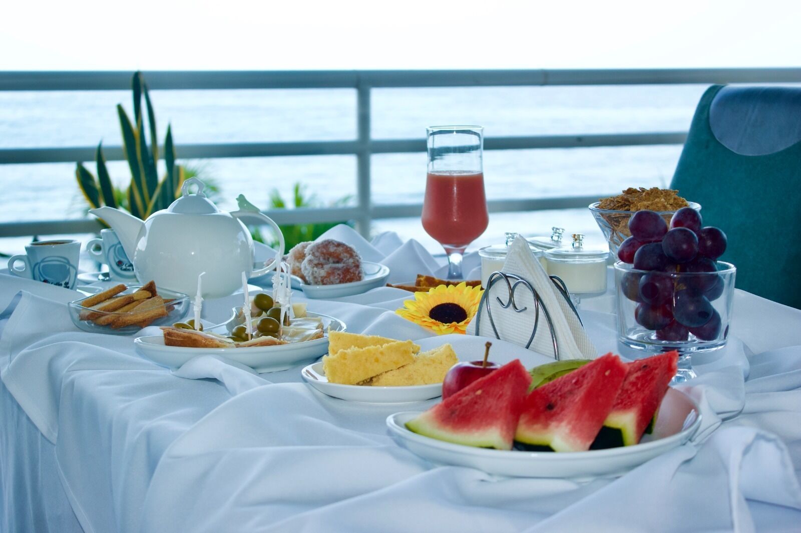 A breakfast table with various food items and a glass of juice, set against a view of the sea.