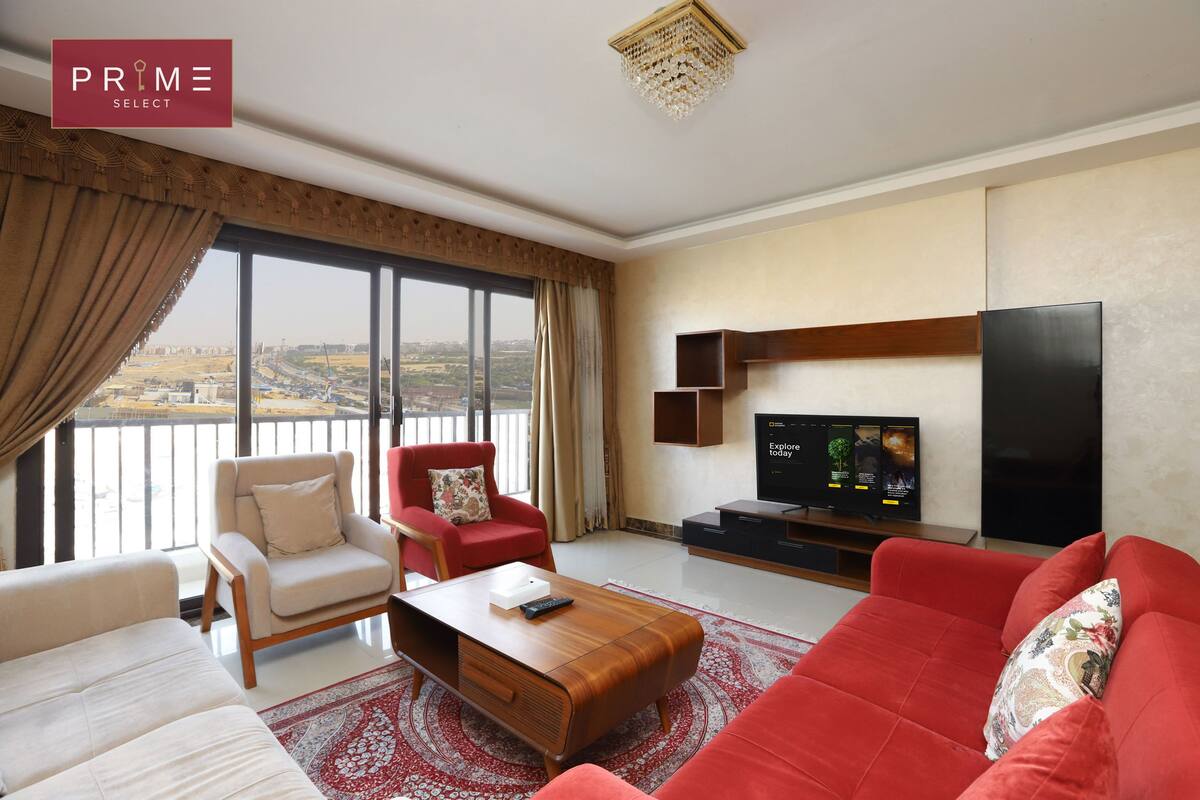 A spacious living room with a large window, a red sofa, a beige armchair, and a wooden coffee table.