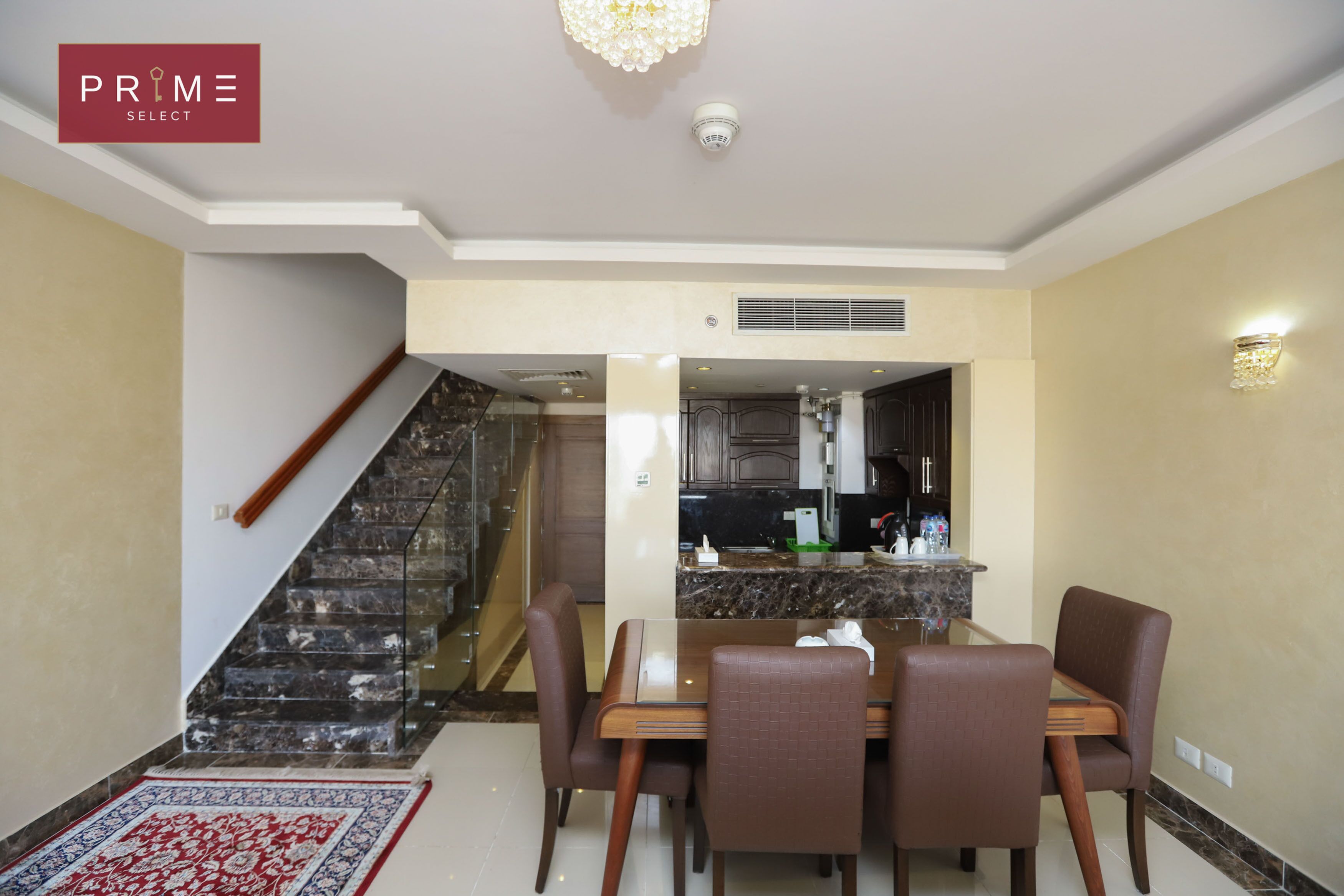 Un comedor con mesa de vidrio y sillas marrones, una escalera y una cocina visible a través de un arco.