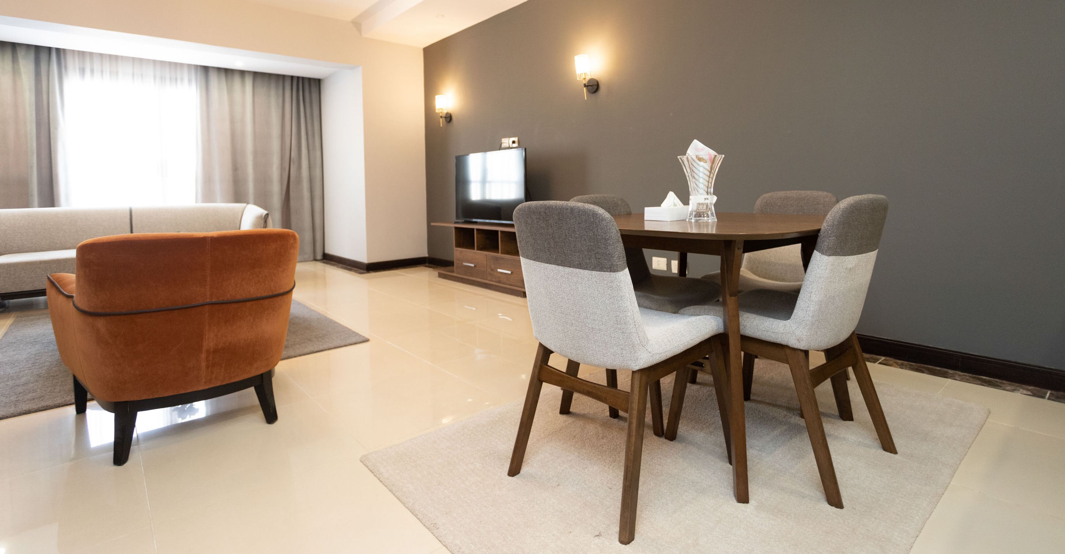 A modern living room with a sofa, a dining table with four chairs, a flat-screen TV, and a vase with a magazine.
