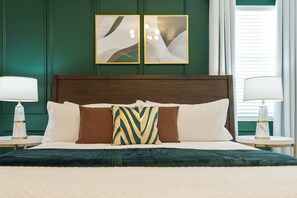 Un lit bien fait, avec une tête de lit vert foncé, deux lampes de chevet et deux tableaux abstraits accrochés au mur.