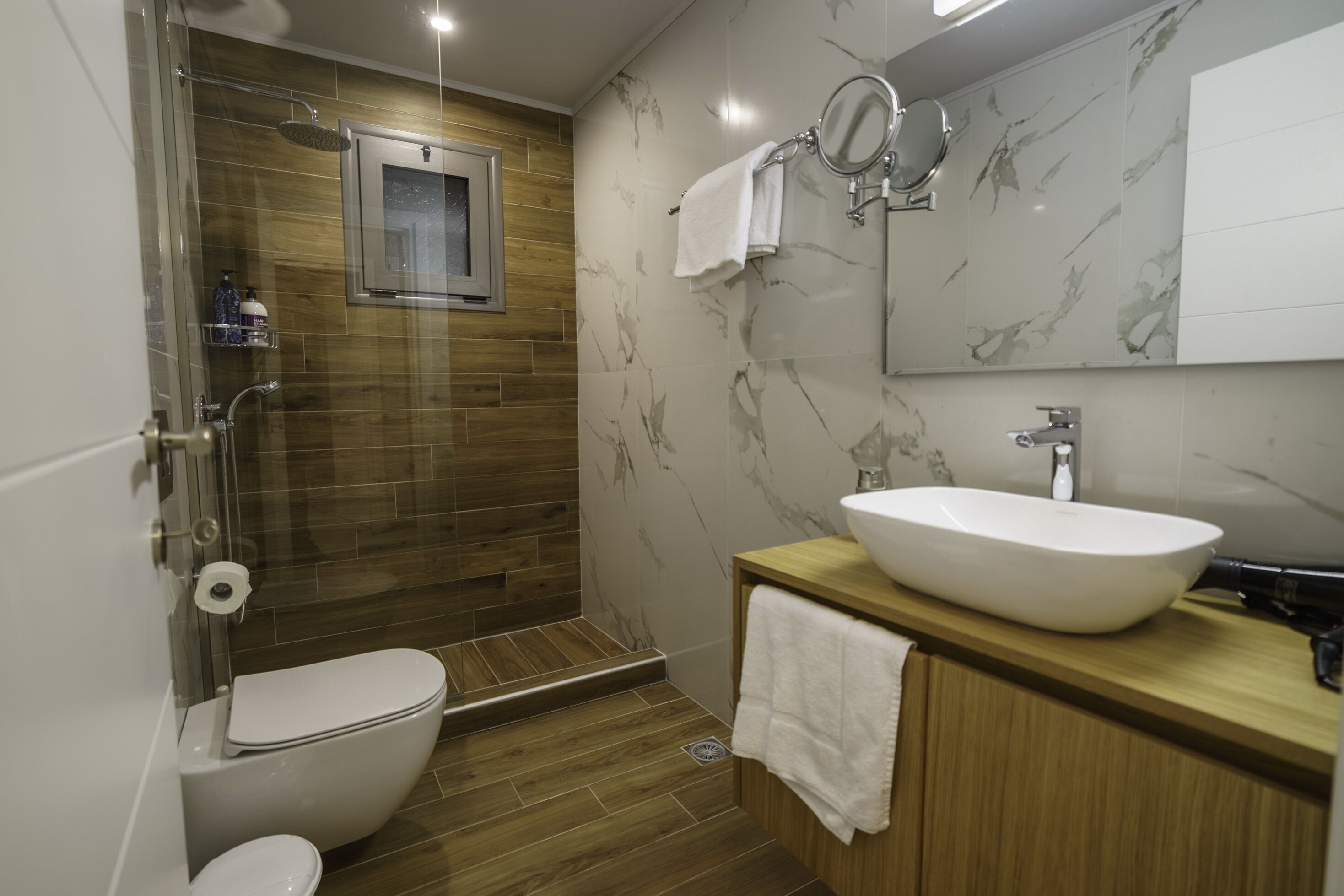 Un baño moderno con ducha de vidrio, tocador de madera con lavamanos blanco y un inodoro.