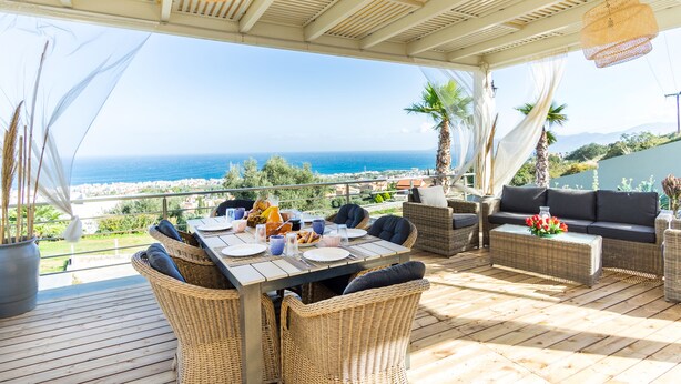 An outdoor dining area with wicker furniture, a table set for a meal, and a view of the ocean. - Royal Hill Villa (Hersonissos)