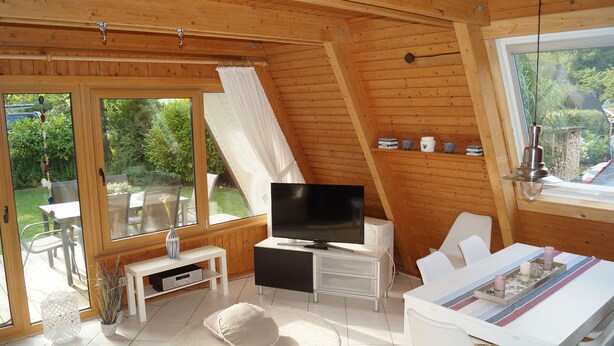 Una habitación moderna y bien iluminada con una televisión grande, muebles blancos y vistas a un jardín a través de amplios ventanales.