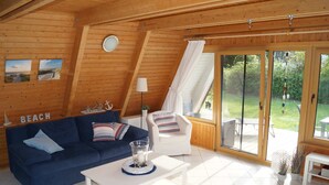Una cabaña acogedora con interior de madera, un sofá azul, un sillón blanco, una mesita y un ventanal corredizo que da a un patio exterior.