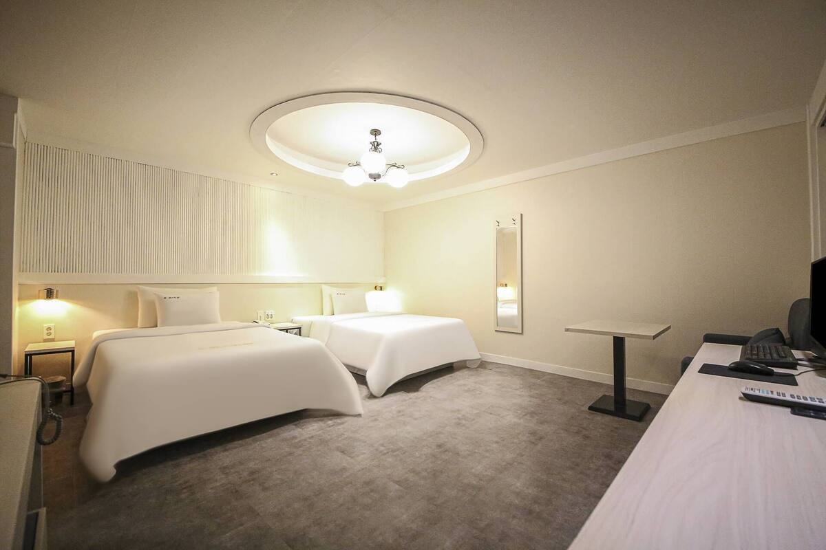 A hotel room with two beds, a desk with a computer, a mirror, and a ceiling light.