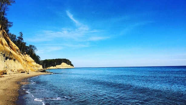 A coastal beach with clear blue waters, sandy shore, and a cliff with vegetation.
