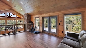 A spacious room with a wooden floor, a leather sofa, a stone fireplace, and large windows offering a view of the outdoors. - Chalet Village | Mountain Views | Resort Pools (Gatlinburg)