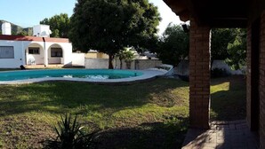Un área de piscina con césped, un edificio blanco con arcos y un árbol grande.