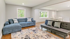 A spacious living room with a grey sofa, a dark grey sofa, and a patterned rug. There are two windows providing natural light. - Hampton Bays Retreat: Walk to Squires Beach! (Hampton Bays)