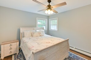 Une chambre à coucher avec un lit en bois, une table de chevet, un ventilateur de plafond et deux fenêtres.
