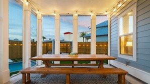 Un porche techado con una mesa y bancos de madera, luces colgantes y vistas a una piscina y casas vecinas.
