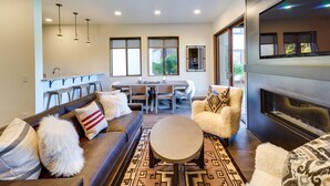 A modern living room with a sofa, armchairs, a coffee table, and a fireplace. There is a dining area with a table and chairs, and large windows providing natural light. - Sun Valley Condo w/ Furnished Patio: Near Skiing! (Sun Valley)