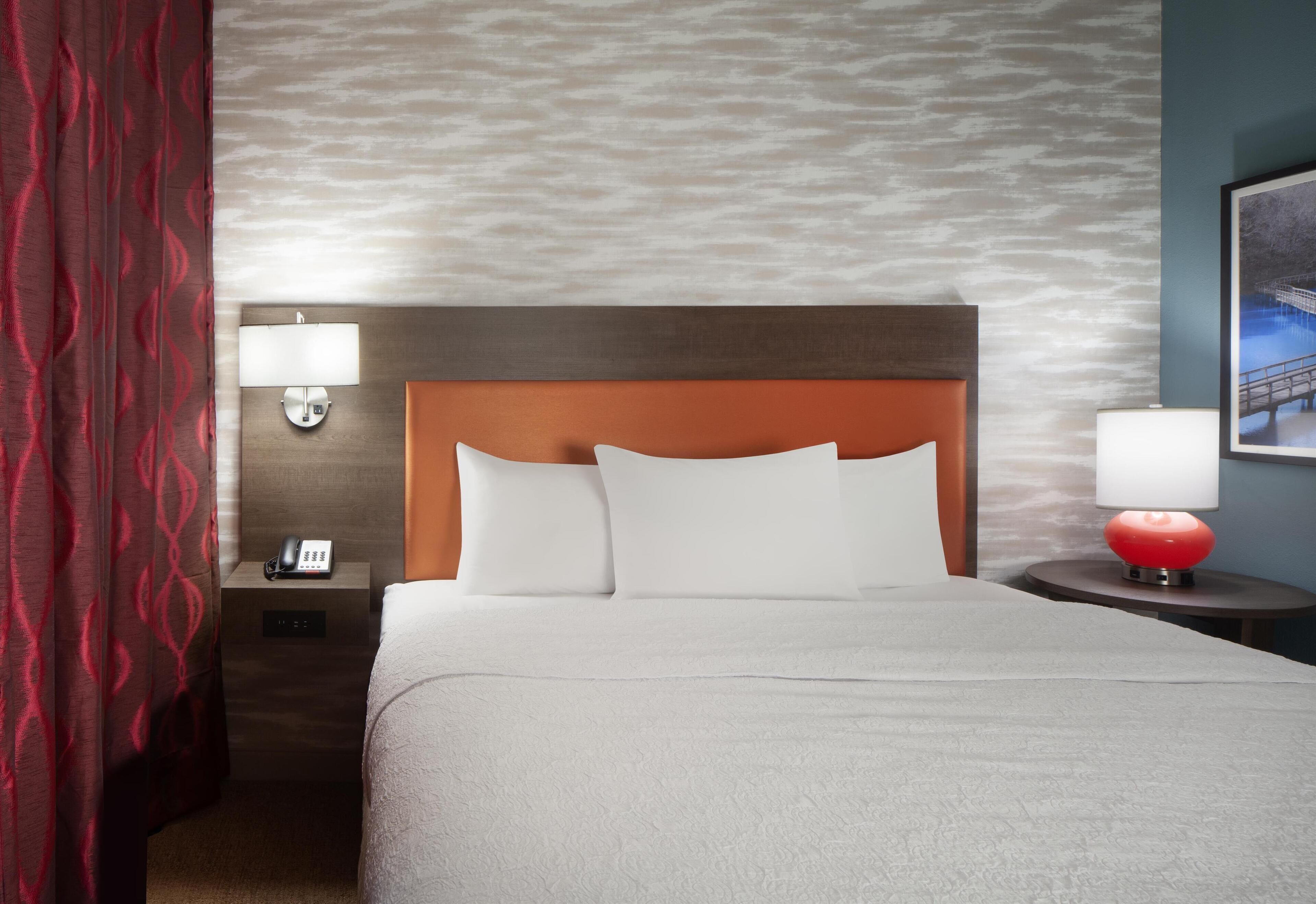 A hotel room with a bed, a nightstand with a lamp, a telephone, and a red lamp on a side table.