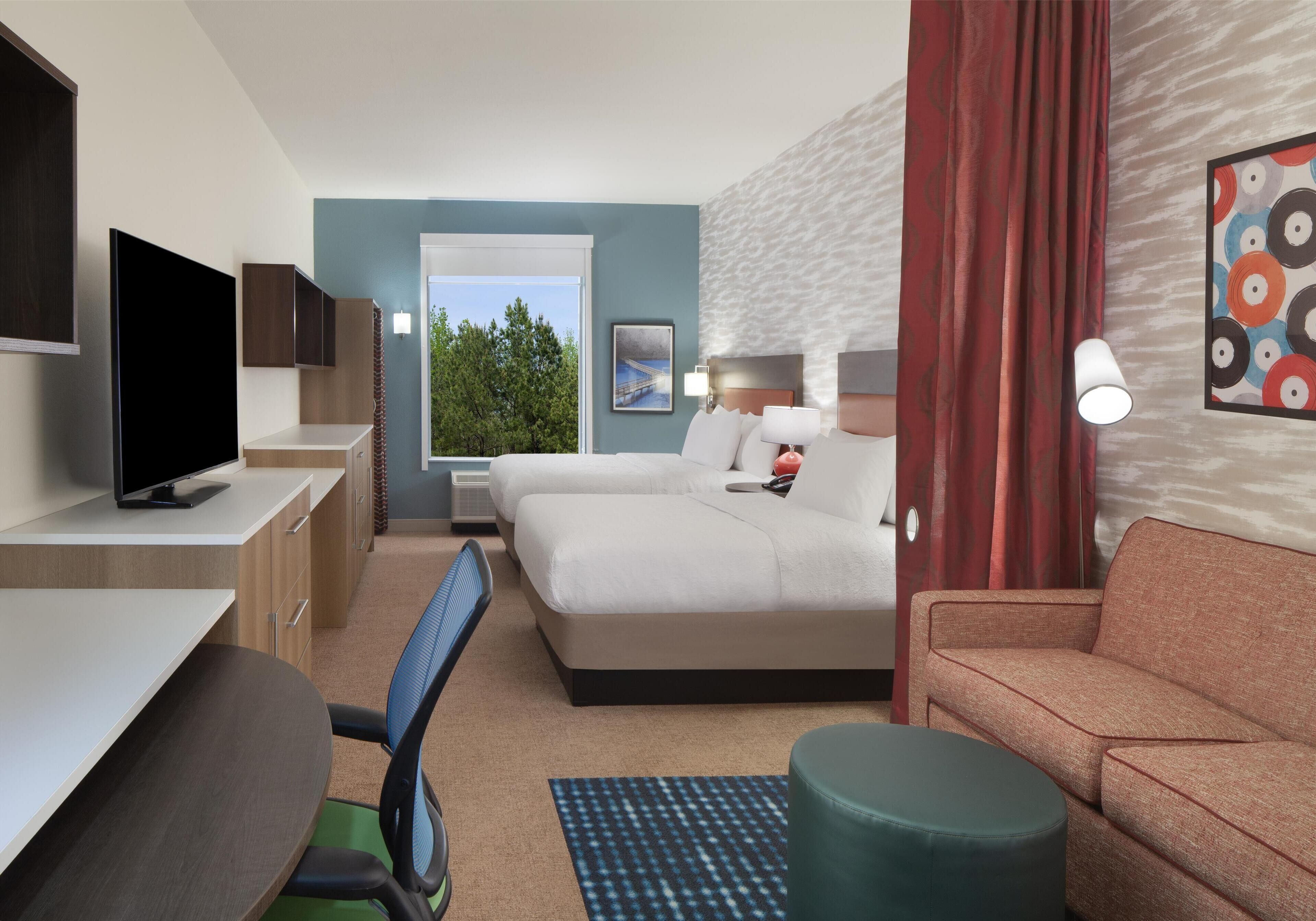 Habitación de hotel con cama, escritorio, televisor, ventana con vista a árboles y cortina roja.