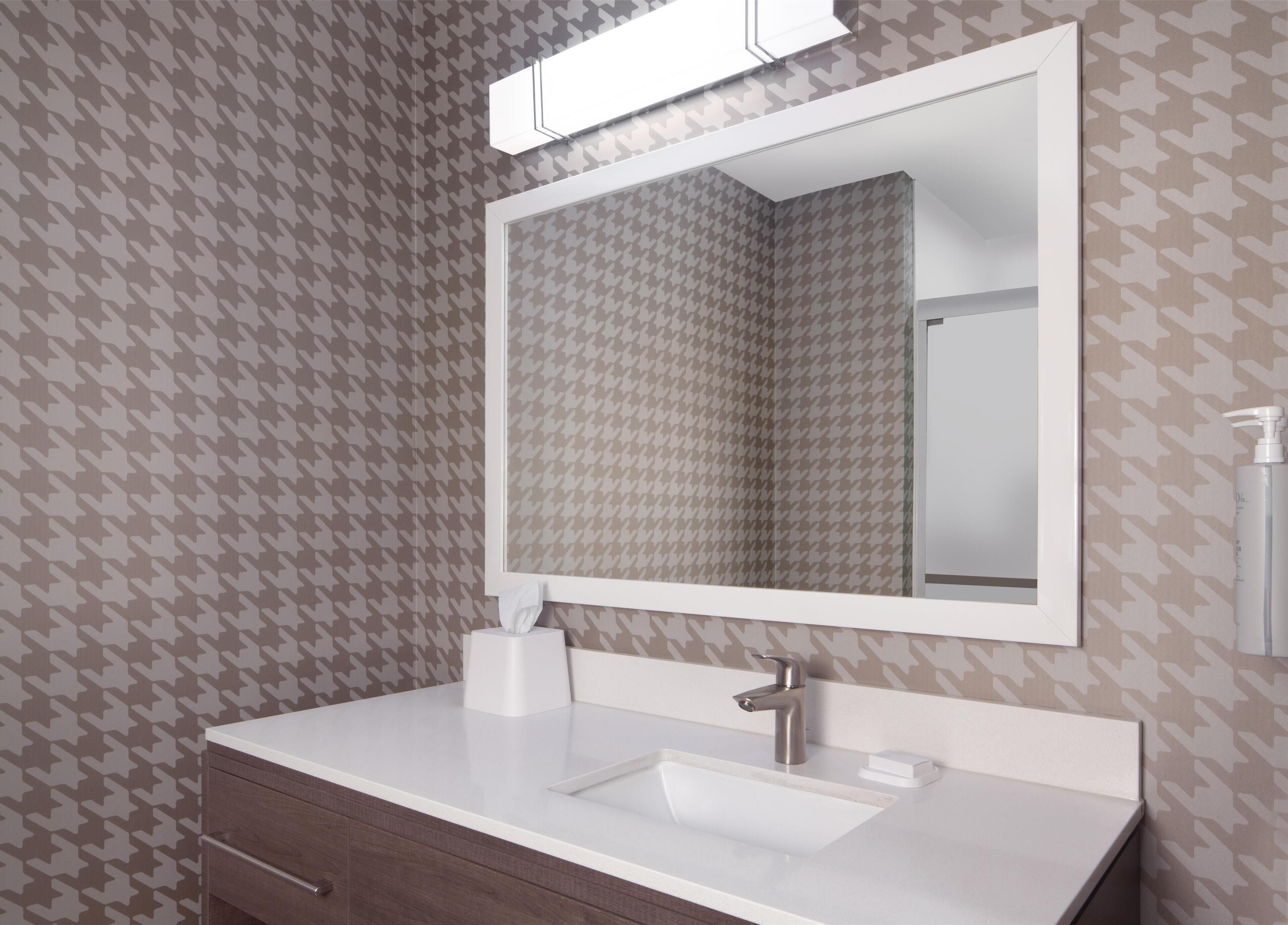 Un baño con papel tapiz estampado con diseño de piedritas, un lavamanos blanco con grifo plateado, un espejo sobre el lavamanos y una caja de pañuelos en la encimera.