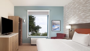 A hotel room with a bed, a TV, a desk, a chair, a window with a view of trees, and a wall with a textured pattern. - Home2 Suites By Hilton Jackson (Jackson)