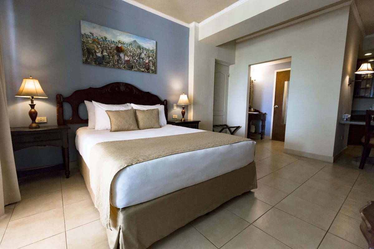 A hotel room with a large bed, two bedside tables with lamps, a painting on the wall, and a door leading to another room.