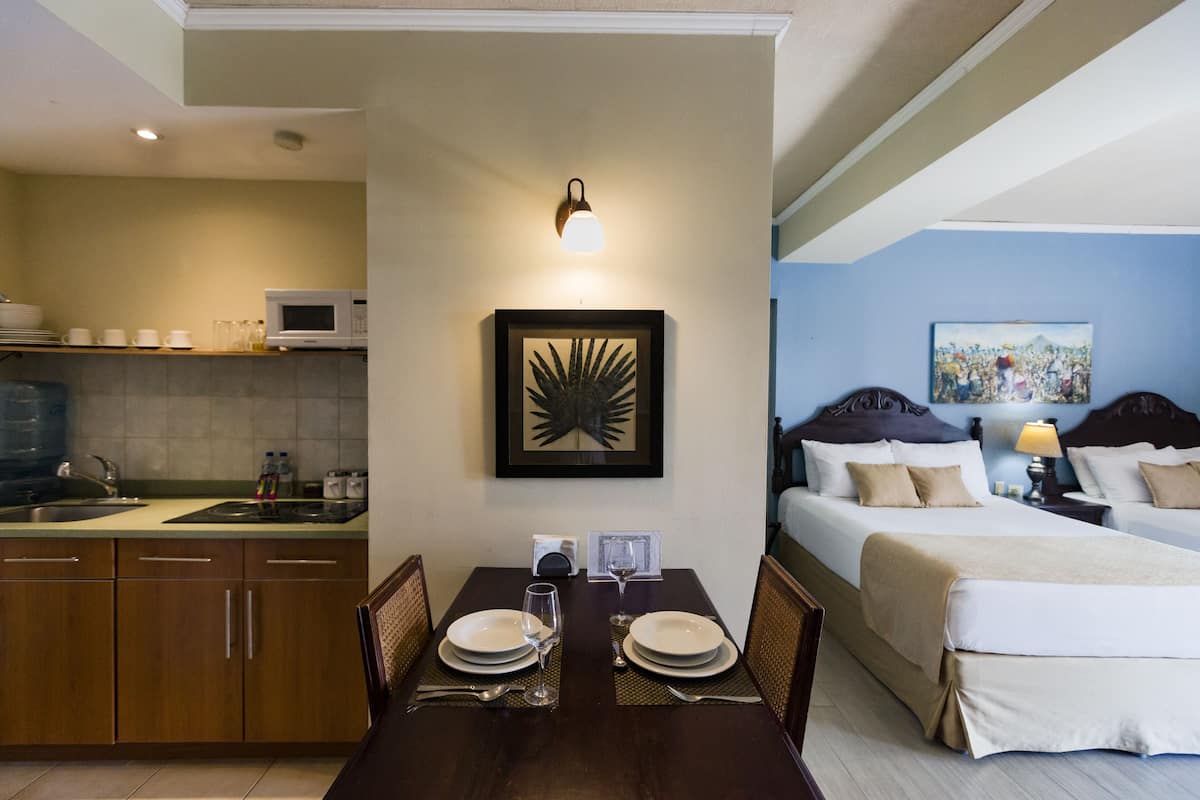 A hotel room with a dining area, a bed with a headboard, a painting on the wall, and a nightstand with a lamp.