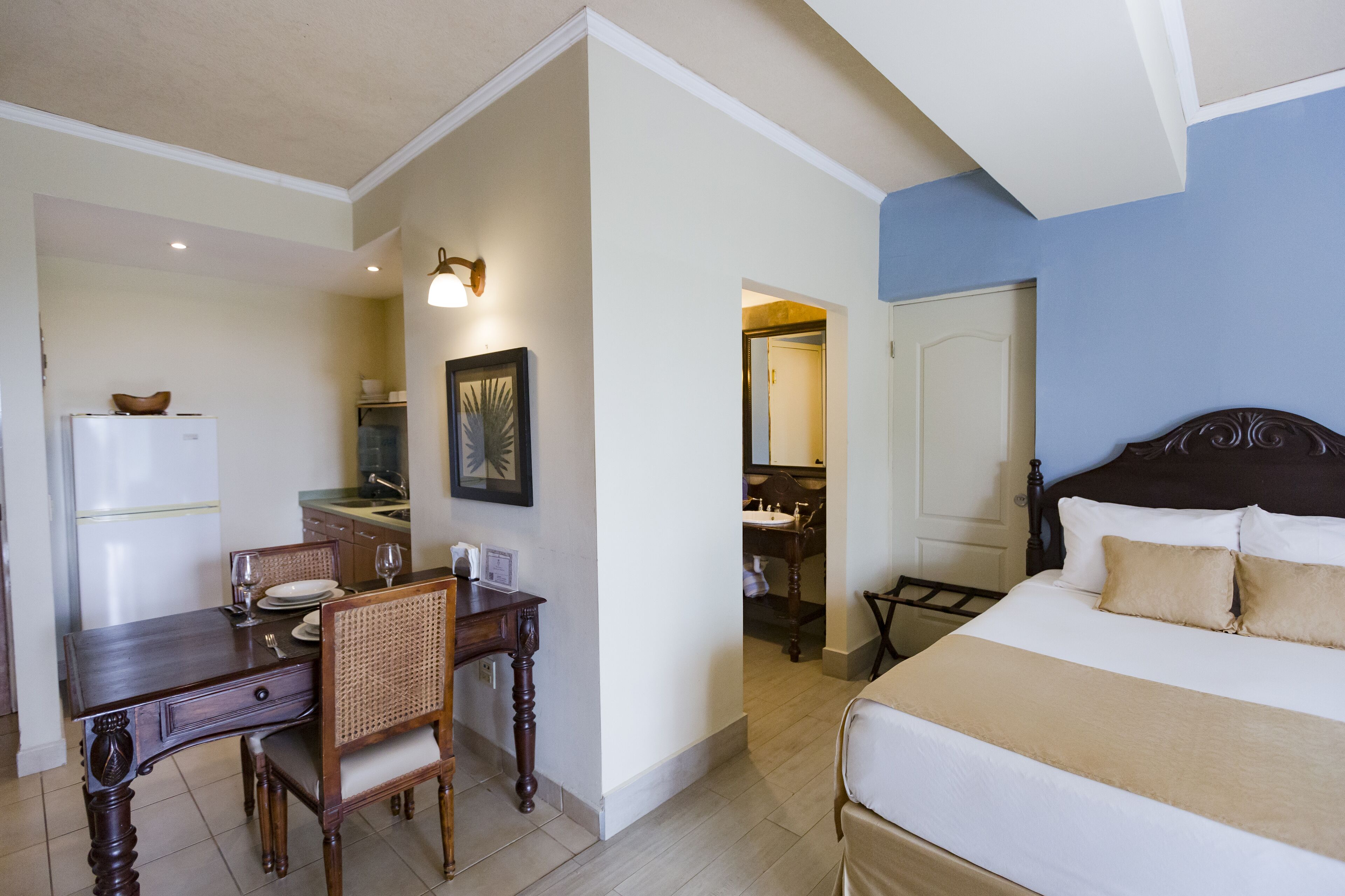 A hotel room with a bed, a dining table with chairs, a refrigerator, a sink, and a mirror.