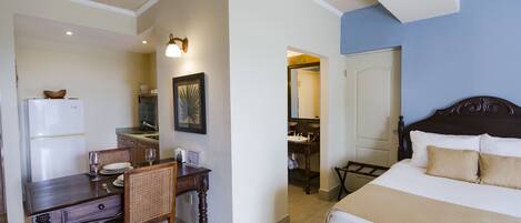 A hotel room with a bed, a dining table with chairs, a refrigerator, a sink, and a mirror.