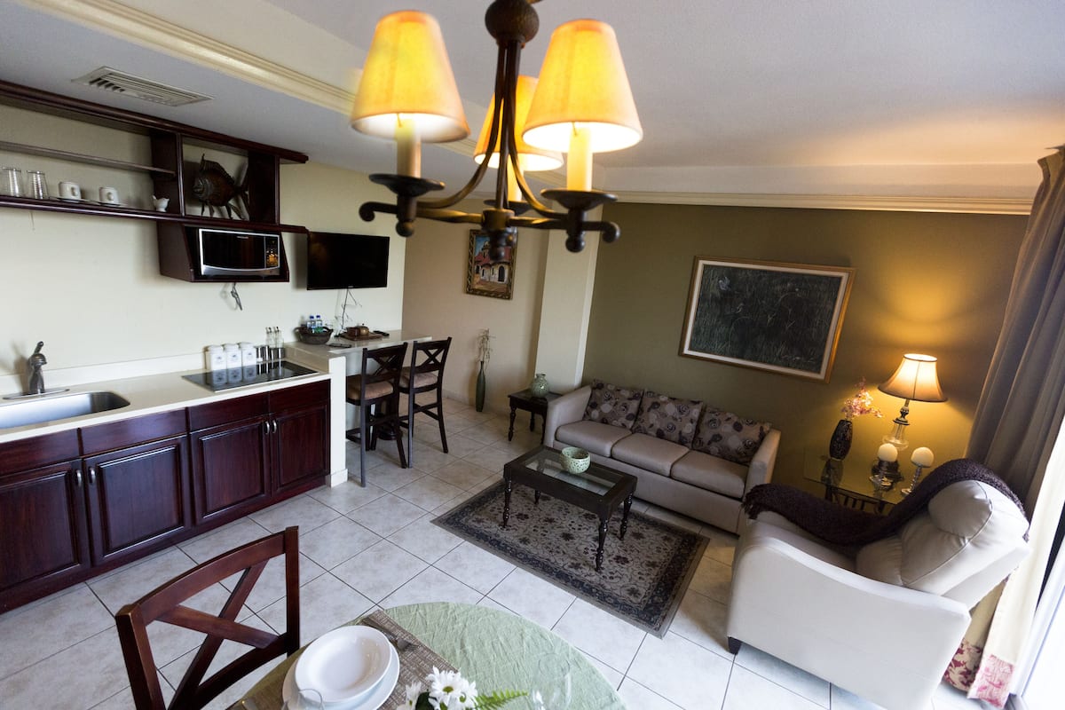 A living room with a sofa, a coffee table, a dining table with chairs, a kitchen area with cabinets and a microwave, and a chandelier.