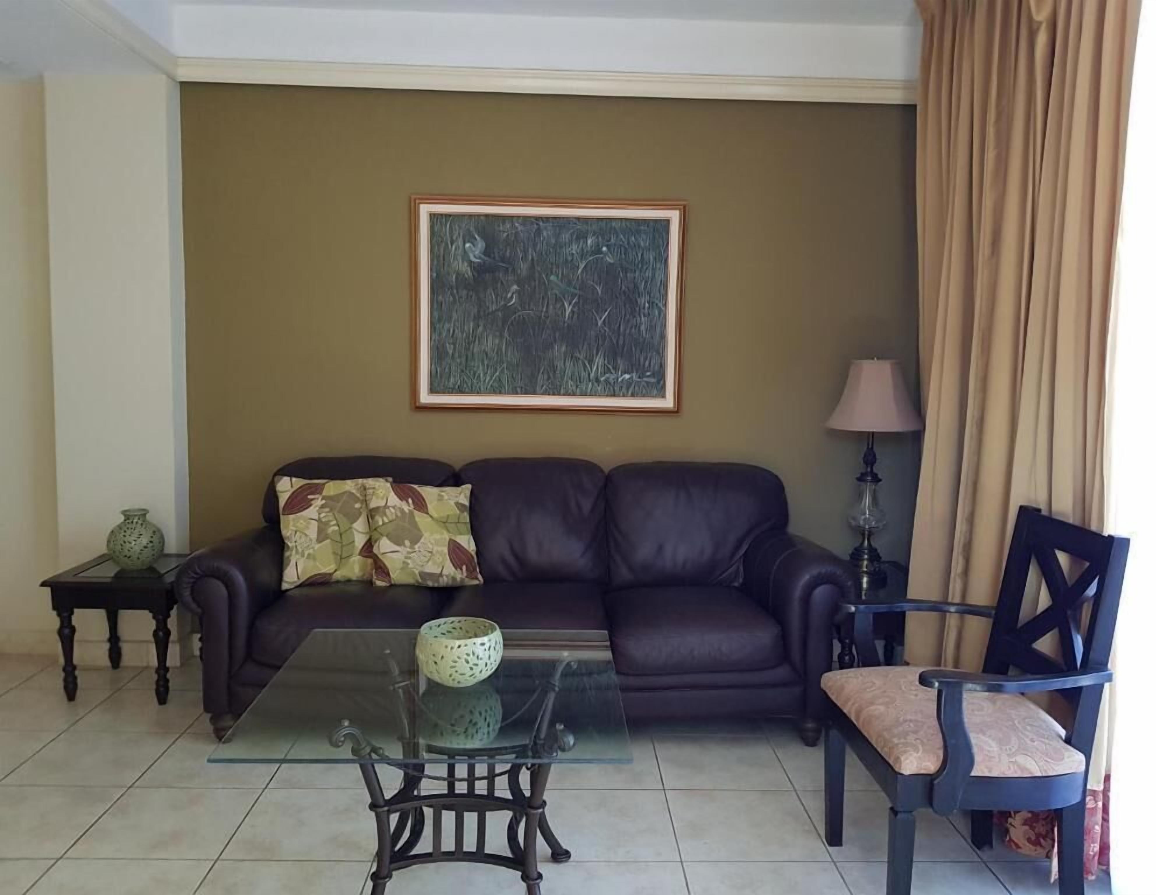 A living room with a dark leather sofa, a glass coffee table, a framed abstract painting, a lamp, and a chair.