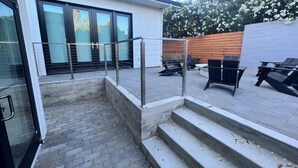 Eine moderne Außenpatio mit einer Betontreppe, die zu einem Glastür-Eingang führt, umgeben von Gartenmöbeln und einem Holzzaun.